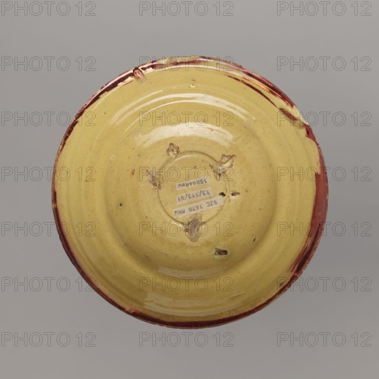 Plate, Manises ceramic center; ca. 1400-,  manufacturer author, mirrors decoration, birds decorative motif, plant decorations. Plate - a type of semi-deep bowl, with a thickened rim. Painted with red mirror in the motif of a bird with spread wings in the center, with volute plant motifs all around. Traces of supports used during firing on the face. 16th/17th century, Spain; Manises. Vessel; height 4.5 cm, width 20.0 cm, A round ceramic dish viewed from above with a glossy yellow interior and a rim showing chipped red and brown glaze, several small rough patches and areas of wear on the surface, and a few spots where the glaze is missing, set against a neutral gray background.