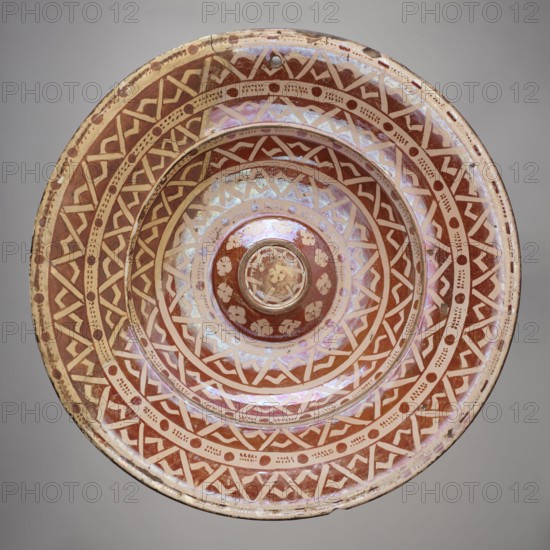 Platter, Manises ca. 1400–, Round ceramic platter with a wide collar and a bulging central cup-shaped bottom. Decorated with four concentric bands of varying widths filled with a geometric ornament resembling a braid composed of two intersecting zigzags; between them are white bands with an ornament of circles and dots. Around the central disc is a trail of four-leaf rosettes; a rosette in the center of the bottom. On the bottom: a wavy star, a spiral, and two rows of oval leaves at the edge. Geometric-floral ornament; 16th century, Spain Manises. Vessel; height 5 cm, width 40 cm., A round ceramic bowl with concentric decorative bands featuring geometric patterns and dot motifs in shades of reddish-brown and cream, showing an iridescent sheen with pale pink and purple highlights; the bowl has a raised central boss and visible wear with small chips and surface scratches; the background is a neutral light gray.