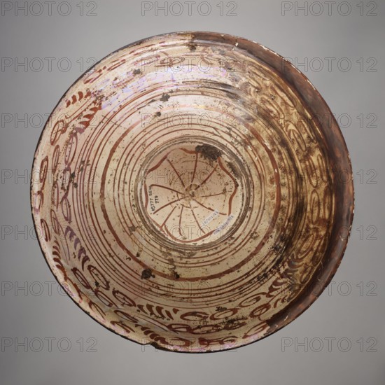 Platter, Manises ceramic center; ca. 1400–, maker role/classification, mirrors decoration; geometric-floral ornament; rosettes. Round platter with a wide collar and a bulging center of the cup-shaped bottom. Decorated with four concentric bands of varying widths, filled with a geometric ornament resembling a braid composed of two intersecting zigzags. Between them are white bands with an ornament of circles and dots. Around the central disc is a trail of 4-leaf rosettes. In the center of the bottom is a rosette. On the bottom: a wavy star, a spiral and two rows of oval leaves at the edge. 16th century, Spain; Manises. Vessel. Height 5 cm, width 40 cm., A round shallow dish viewed from above with concentric circular grooves and decorative curved brushwork around the rim, showing a pale cream base color with brown painted patterns and a few areas of darker brown and rust-colored discoloration, along with some iridescent sheen and small dark spots of surface wear.