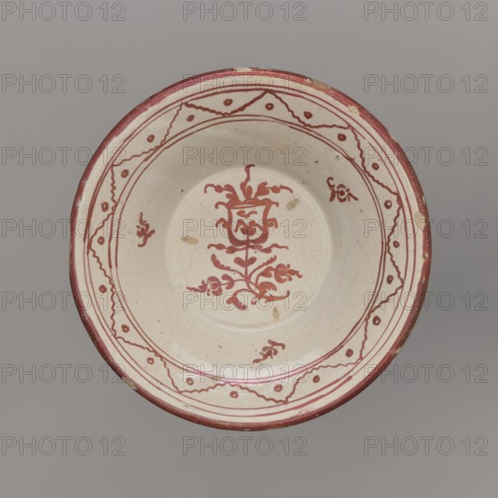 Bowl, unspecified manufacturer; mirrors decoration; geometric-floral ornament; bowl decorated on the collar with a pattern of triangles and small circles; in the mirror a stylized tulip sprig and three small flower patterns; 18th century, Spain; vessel; height 4 cm, width 18 cm., A round shallow ceramic dish with a beige base color and red decorative painting, showing a central floral motif in red with curling stems and leaves, a few small red ornamental marks around the inner rim, and a red patterned border with wavy lines and dots near the outer edge; the rim itself is painted red with a few small areas of wear revealing the lighter base, and the background behind the dish is a uniform gray.