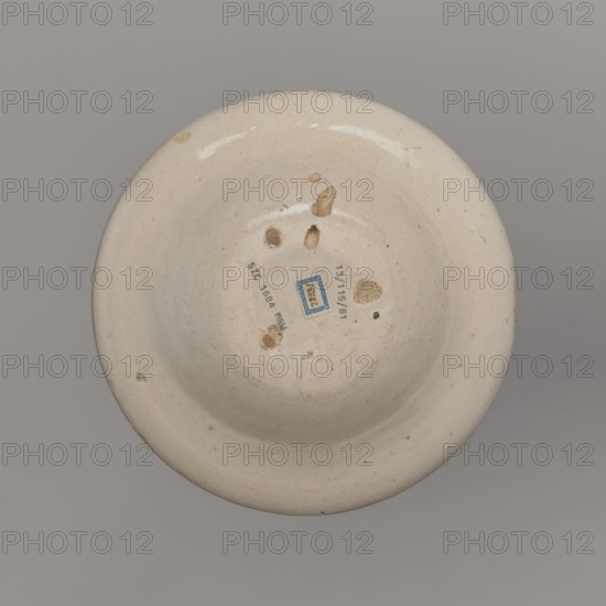 Bowl, unspecified manufacture; mirrors decoration; ; geometric-floral ornament; bowl decorated on the collar with a pattern of triangles and small circles; in the mirror a stylized tulip sprig and three small flower patterns; 18th century, Spain; vessel; height 4 cm, width 18 cm, A round shallow bowl viewed from above with a creamy off-white glazed surface showing a few irregular brownish chips and spots on the interior and rim, a group of small circular and oval indentations toward the center, a small rectangular blue patch near the middle, and a plain gray background surrounding the object.