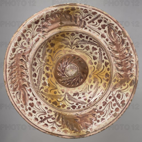 Round platter, profiled with a slightly convex center of the bowl-shaped bottom, with a cylindrical rim. Painted with floral and plant motifs. Four fleshy leafy motifs are hand-embossed on the collar. The remaining surface of the collar is covered with plant decoration. In the mirror, on a floral wallpaper, is a four-petaled flower motif made of leaves, with a spiral rosette at the bottom. Decoration made with red mirror. Platter, Manises ceramic center; ca. 1400-, 16th/17th century, Spain; Manises. Vessel. Height 10 cm, width 50 cm., A shallow decorative bowl seen from above with a central raised spiral element and a wide rim, featuring embossed leaf-like forms and painted floral and foliate motifs; colors visible include shades of gold, beige, brown, pink, and reddish-brown, with highlights of pale cream and subtle greenish-gray on the background.