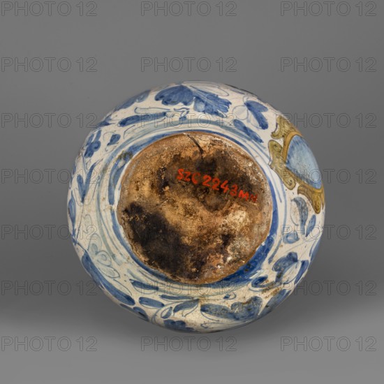 Bottle. Spherical bottle, narrowed at the top with a pressed rim, the foot slightly widening at the base. White ceramic painted with cobalt of varying saturation, with loosely scattered plant motifs resembling oak leaves, flowers, and acorns. On the front of the body, a coat of arms cartouche under a banderole no inscription. The blue-and-white leaf decoration shows Venetian influence. Maker: Montelupo pottery workshop Montelupo, Italy, 2nd half 16th century. Height 24.5 cm; width 18.0 cm., A round ceramic object with a worn, discolored central area showing rough brown and dark brown patches, surrounded by a glazed surface decorated with blue floral and leaf-like brushstrokes on a white background, and a small area of pale gold and light blue ornamentation along the rim; the overall scene sits against a smooth gray backdrop.