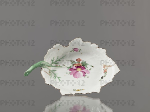 Leaf-shaped bowl. Makers: Francis Gardner porcelain factory 1766–1917 and Wierbilki/near Moscow porcelain factory from 1766. Leaf-shaped bowl on a ring-shaped foot. Rims cut in the shape of leaves with a triangular rim; opposite side a handle in the form of a green stem with veining. Decoration: at the bottom a bouquet with a large purple-pink flower and three small floral motifs. Condition: four cracks extending from the rim, glaze abrasions. Date: 1775–1800. Place: Russia, Wierbilki. Function: dish. Dimensions: height 9.5 cm; width 12.0 cm. Provenance: purchase., A scalloped porcelain dish shaped like a leaf with a green stem-like handle, decorated with painted floral motifs including a prominent pink flower with yellow center and smaller orange and yellow blossoms, set against a white surface with a gilded rim; the background is gray and the dish casts a soft reflection below.