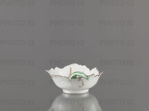 Leaf-shaped bowl, Wierbilki porcelain factory; 1766-, maker: Gardner, Francis porcelain factory; 1766–1917. Leaf-shaped bowl on a ring-shaped foot. Rims cut in the shape of leaves with a triangular rim; opposite side a handle in the form of a green stem with veining. At the bottom is a bouquet with a large purple-pink flower; around it three small floral motifs. Four cracks extending from the rim, glaze abrasions. 1775–1800, Russia. Dish; height 9.5 cm, width 12.0 cm., A small white scalloped bowl with a glossy finish sits centered against a smooth gray background, its rim edged with a thin brownish trim and showing a few brownish spots on the exterior; a small green curved leaf-like ornament rests on the inner rim, and the bowl casts a soft reflection on the surface beneath it, with visible colors including white, gray, brown, and green.