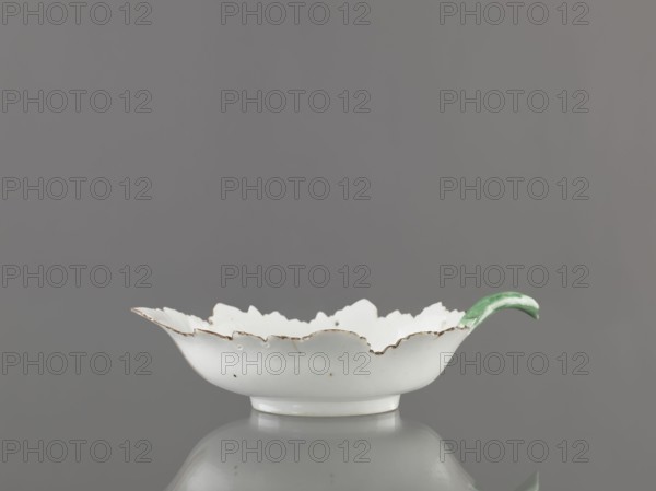 Leaf-shaped bowl, leaf-shaped bowl on a ring-shaped foot. Rims cut in the shape of leaves, triangular rim, on the opposite side a handle in the form of a green stem with veining. At the bottom is a bouquet with a large purple-pink flower, around it three small floral motifs. Four cracks extending from the rim, glaze abrasions. Porcelain, 1775–1800, Russia, Wierbilki; height 9.5 cm, width 12.0 cm, A shallow white ceramic dish with a scalloped rim showing worn brown edging and small speckles sits on a reflective surface against a gray background, with a curved green handle attached to one side.