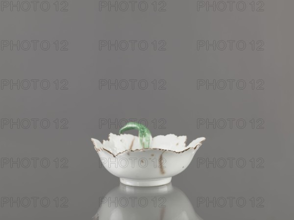Leaf-shaped bowl. Makers: Francis Gardner porcelain factory 1766–1917 and Wierbilki/near Moscow porcelain factory founded 1766. Leaf-shaped bowl on a ring-shaped foot; rims cut in the shape of leaves with a triangular rim; opposite side a handle in the form of a green stem with veining. Decoration: at the bottom a bouquet with a large purple-pink flower and three small floral motifs. Condition: four cracks extending from the rim, glaze abrasions. Date: 1775–1800. Place: Russia, Wierbilki. Function/type: dish. Dimensions: height 9.5 cm; width 12.0 cm., A small white ceramic bowl with a scalloped, irregular rim and worn brownish edges sits centered on a reflective gray surface against a smooth gray background, and a curved green glass handle or decorative element rises from the bowl's center; the bowl shows a few brownish stains and slight discoloration, and the colors visible are white, gray, green, and brown.
