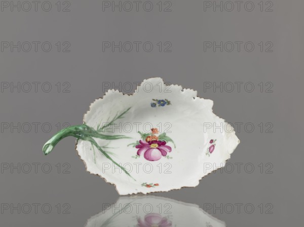 Leaf-shaped bowl, Wierbilki/near Moscow porcelain factory; est. 1766. Maker: Gardner, Francis porcelain manufactory; 1766–1917. Leaf-shaped bowl on a ring-shaped foot. Rims cut in the shape of leaves with a triangular rim; opposite side a handle in the form of a green stem with veining. At the bottom is a bouquet with a large purple-pink flower, surrounded by three small floral motifs. Reconstructed tip of the spout; abrasions of the glaze. Date: 1775–1800. Place: Wierbilki, Russia. Vessel; height 9.5 cm, width 12.0 cm., A shallow, irregularly edged white dish with a scalloped rim and a small green handle shaped like a stem, decorated with painted floral motifs including a central pink and purple flower with green leaves, smaller blue and orange flowers, and green brushstroke accents; the rim has a thin gold-colored trim and the dish sits on a reflective gray surface with a solid gray background.