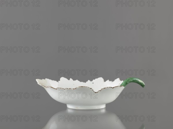 Leaf-shaped bowl, Wierbilki/near Moscow porcelain factory 1766–, attributed to Gardner, Francis porcelain manufactory, 1766–1917. Leaf-shaped bowl on a ring-shaped foot. Rims cut in the shape of leaves with a triangular rim; opposite side a handle in the form of a green stem with veining. At the bottom is a bouquet with a large purple-pink flower and three small floral motifs. Reconstructed tip of the spout; abrasions of the glaze. 1775–1800, Wierbilki, Russia. Vessel; height 9.5 cm, width 12.0 cm., A shallow white ceramic bowl with a scalloped rim that has uneven, slightly chipped edges and a decorative green curved handle attached to one side, set against a neutral gray background with a soft reflection beneath; visible colors include white, green, gray, and small areas of brownish discoloration along the rim.
