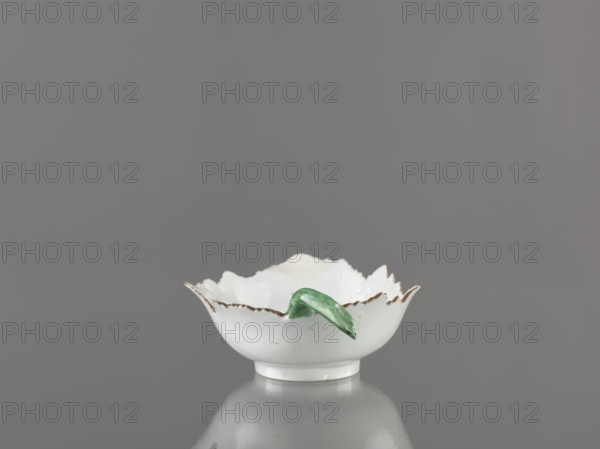 Leaf-shaped bowl, Wierbilki/near Moscow porcelain factory; 1766–, Gardner, Francis porcelain manufactory; 1766–1917; purchase provenance. Leaves; bowl on a ring-shaped foot. Leaf-shaped bowl on a ring-shaped foot with rims cut in the shape of leaves and a triangular rim; opposite side a handle in the form of a green stem with veining. At the bottom is a bouquet with a large purple-pink flower surrounded by three small floral motifs. Reconstructed tip of the spout; abrasions of the glaze. 1775–1800, Russia. Vessel; height 9.5 cm, width 12.0 cm., A small white ceramic bowl with a scalloped rim and a few worn brown edges sits centered on a reflective surface, with a green ceramic leaf resting on its rim; the background is a uniform medium gray and the visible colors are white, green, brown, gray.