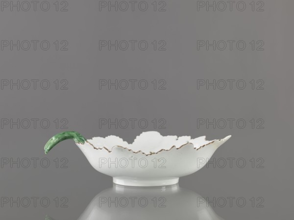 Leaf-shaped bowl, Wierbilki/near Moscow porcelain factory est. 1766. Maker: Gardner, Francis porcelain manufactory; 1766–1917. Leaf-shaped bowl on a ring-shaped foot. Rims cut in the shape of leaves with a triangular rim; opposite side a handle in the form of a green stem with veining. At the bottom a bouquet with a large purple-pink flower and three small floral motifs. Reconstructed tip of the spout; abrasions of the glaze. Date: 1775–1800. Place: Wierbilki, Russia. Vessel. Height 9.5 cm; width 12.0 cm., A shallow white ceramic dish with a scalloped rim showing a slightly irregular edge and a narrow rounded foot, sitting on a reflective gray surface against a smooth gray background; the rim has subtle brownish accents and one side of the dish features a curved handle glazed in green.