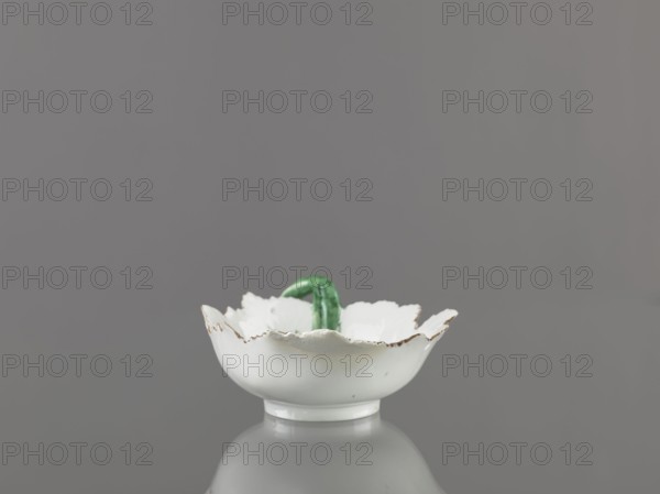 Leaf-shaped bowl, Wierbilki/near Moscow porcelain factory; 1766- . Leaf-shaped bowl on a ring-shaped foot. Rims cut in the shape of leaves with a triangular rim; on the opposite side a handle in the form of a green stem with veining. At the bottom is a bouquet with a large purple-pink flower, around it three small floral motifs. Reconstructed tip of the spout; abrasions of the glaze. 1775–1800. Russia. Wierbilki. Vessel. Height 9.5 cm; width 12.0 cm., A small white scalloped bowl with a slightly worn rim sits on a reflective gray surface, showing a reflection beneath it; inside the bowl a curved green object is partially visible; the background is a uniform medium gray.