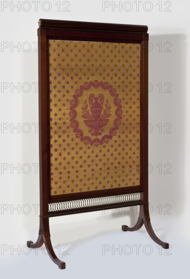 Fireplace screen, origin unknown, Empire style, wattle ornament, plant decoration, rosette ornament, decorative vases. Rectangular mahogany-veneered frame filled with patterned silk fabric, mounted on a pair of bipod supports. Posts and horizontal stiles are angular; the lower stile is an openwork brass strip with a wattle motif. Supports set in cubes are two arched, tapering angular legs ending in angular feet. Fabric stretched on an inner frame inserted from above into grooves of the side posts, enclosed by narrow slats; upper stile topped with a roller added during conservation replacing a non-original simy-profile strip. Brass rosettes on the sides of the shaft. Fabric: yellow plain-weave ground with a red satin pattern originally reversed color arrangement: central oval field with a vase on a pedestal framed by twigs; remaining field filled with small rosettes. Reverse upholstered with yellow silk fabric previously cherry-red linen canvas. A flat steel spring inside one side frame...