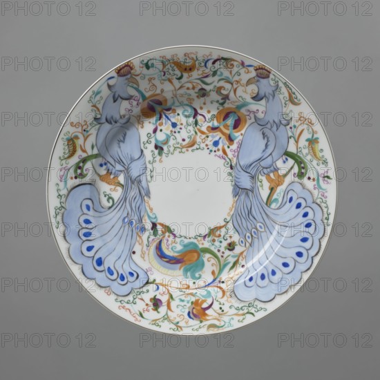 Plate, porcelain; factory named after Mikhail Vasilyevich Lomonosov 1918–. Plate with colorful decoration spread in a wide band around the edge — two stylized blue peacocks between plant scrolls and orchid flowers; center left undecorated. Dish; porcelain; 1920; Leningrad, Russia. Height 4 cm; diameter 24.8 cm., A round decorative plate with a white background featuring a symmetrical design of two stylized birds facing each other, each rendered in pale blue with long flowing tail feathers that have darker blue teardrop-shaped accents, and golden beaks topped with small crown-like crests; the central rim area around a blank circular center is filled with an ornate pattern of curling vines and floral motifs in green, gold, orange, purple, teal, and blue, with the decorative elements forming a balanced, mirrored composition; the plate has a thin rim in metallic gold and is shown against a neutral gray background.