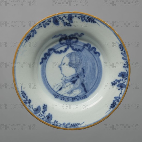 Plate, Delft ca. 1600–, De Porcelejne Bijl faience manufactory; 1657–1803, busts, gift, princes, portraits of monarchs, plant decorations, letters. In the mirror, a chiaroscuro portrait of a man in left profile, painted in cobalt, with a ribbon and the letters WP 5 PVOR. The portrait depicts Prince William V 1748–1806, ruler of Orange. The inscription can be translated as Willem Rex Prins Van ORanie. Similar plates were produced after 1787 in Delft. 2nd half of the 18th century, Netherlands, Delft. Dish; height 5 cm, width 23.5 cm., A round ceramic dish with a pale off-white background and a yellowish rim, decorated in blue with a central circular portrait of a person in profile surrounded by a decorative ribbon-like frame, blue floral and leafy motifs around the inner edge, and blue shading and detailing throughout.