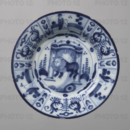 Plate, De Porceleyne Claeuw faience manufacturer; 1658–1840 manufacturer, animals, lions, plant decorations, chinoiserie. Semi-deep plate, quite thick-walled with a collar thinner at the edge. On a light blue background, decoration in dark cobalt. At the bottom, between two arbor posts flanked by schematically outlined bushes, a standing lion with a human face. The collar features six trapezoidal segments separated by narrow vertical fields with floral decoration, three of which depict a seated male figure, the other three arranged alternately with a schematic, symmetrical volute-floral composition. The lion at the bottom likely represents the Batavian Lion, symbolizing the House of Orange. 18th century, Netherlands, Delft; vessel; height 3.0 cm, width 23.2 cm., A round ceramic plate with a white base and blue painted decoration, showing a central scene of a stylized animal with a mane standing beneath an architectural element with columns, surrounded by curved brushstrokes and decorat...