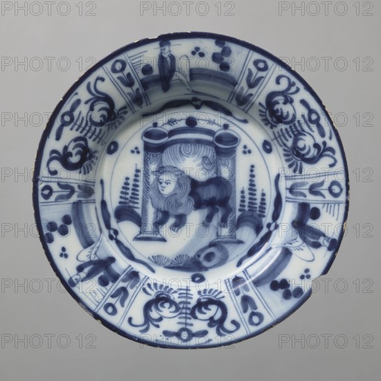 Plate, Lambertus Sanderus 1764-1806 author; De Porceleyne Claeuw faience manufacturer; 1658-1840 manufacturer. Semi-deep faience plate, quite thick-walled with a collar thinner at the edge. On a light blue background, decoration in dark cobalt. At the bottom, between two arbor posts flanked by schematically outlined bushes, a standing lion with a human face. The collar features six trapezoidal segments separated by narrow, vertical fields with floral decoration, three of which depict a seated male figure, the other three arranged alternately with a schematic, symmetrical volute-floral composition. The lion at the bottom likely represents the Batavian Lion, symbolizing the House of Orange. 19th century, Netherlands, Delft. Vessel: height 3.0 cm, width 23.2 cm., A round ceramic plate with blue and white decoration showing a central scene of an animal with a mane standing beneath a small architectural structure with columns, surrounded by stylized foliage and abstract motifs; the rim is d...