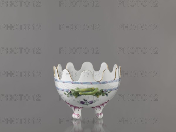 Vernier for glasses, Imperial Porcelain Factory in St. Petersburg c. 1780. Oval vernier set on two volute-shaped arches with a serrated edge; below a strip of relief basketwork framed top and bottom by a ribbon of three rods tied at regular intervals with a diagonal ribbon. Belly with painted colorful floral decoration and gilding. Porcelain vessel, Russia, St. Petersburg. Height 16.5 cm; width 32.5 cm; depth 18.0 cm., A small white porcelain bowl with a scalloped rim trimmed in gold sits centered against a neutral gray background; the bowl has a woven-texture band near its top painted with blue and pink accents, a painted floral motif in purple and green on its body, a raised green decorative handle-like element on the front, and three curled feet with pink shading that reflect faintly on the surface below.