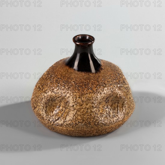Vase, unspecified manufacture; dripstone glaze; Far Eastern-style decorations imitation of Japan; Art Nouveau; bottle-shaped vase with a short, narrow neck and a slightly upturned rim; body flattened in five places; dark brown neck; body covered with a decorative beige-brown glaze with an orange surface resembling a puffball; 19th/20th century, Europe; vessel; height 11.8 cm, width 14 cm., A rounded ceramic vessel with a textured, dimpled surface in shades of brown and tan and a smooth, glossy neck and rim in dark brown to black sits on a pale gray surface casting a soft shadow; the background is a light gray.