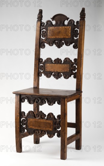 Chair, carved, with a board seat and a high backrest composed of two boards with a wavy, openwork edge, supported by four straight, angular legs, the rear ones merging into slightly rearward-sloping backrest stiles. The legs are fastened with a casement binding with angular joints; the front additionally joined at mid-height by a decorative board shaped analogously to the backrest boards carved with C- and S-shaped acanthus leaves, with single rectangular fields framed with inlaid veining. The side frames are angular and narrow, the front carved with a lower outline repeating the edge of the backrest boards and front legs. The seat edge is profiled in the form of a half-cylinder with a step. The finials of the side backrest frames are in the form of a finial with a volute. Renaissance style; early 17th century; 4th quarter of the 19th century reclaimed/provenance noted;, Southern Germany /Lombardy, origin. Piece of furniture seating piece. Height 121.5 cm, width 51 cm, depth 45.2 cm., ...