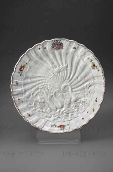 Round platter, Kaendler, Johann Joachim 1706–1775, sculptor; Koenigliche Porzellain Fabrique, Meissen 1710–1763, manufacturer. Swan Service. Deep round platter with a relief of swans, a crane and a heron among reeds; shell relief around the collar; marital coat of arms; polychrome Indian flowers and gilding. 1738–1739. Meissen, Saxony. Porcelain vessel; height 6.5 cm, diameter 29.5 cm., A large white scalloped ceramic plate with a raised relief scene of swans and reeds in the center, surrounded by radiating ridges and a fluted rim, decorated with small painted flowers in red, yellow, purple, blue, green, and orange around the edge, with touches of gold along the rim and a colorful painted crest near the top; the plate rests on a clear plastic stand against a dark gray background.