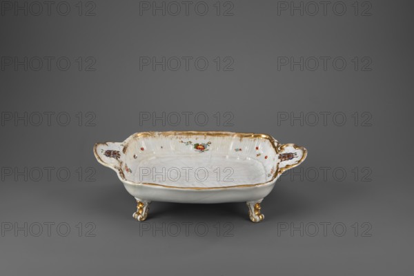 Sweets plate by Johann Joachim Kaendler 1706–1775, modeled with contributions by Johann Friedrich Eberlein 1695–1749, manufactured by Koenigliche Porzellain Fabrique, Meissen Meissen, Saxony, 1740. Rectangular vessel on four volute legs, with shell handles on the short sides, bearing the Brühl marital coat of arms. Bottom relief with swans, a crane and a heron; inner sides with shell relief, Indian flowers, and gilded edge. Height 8.5 cm; width 31.0 cm; depth 20.4 cm., A shallow porcelain dish with ornate handles sits on a neutral gray background; the dish is primarily white with gold edging and gold-accented feet, and it has small floral painted decorations in red and green on the inner rim and handles, with some brownish details on the handles and subtle variations of white on the dish surface.