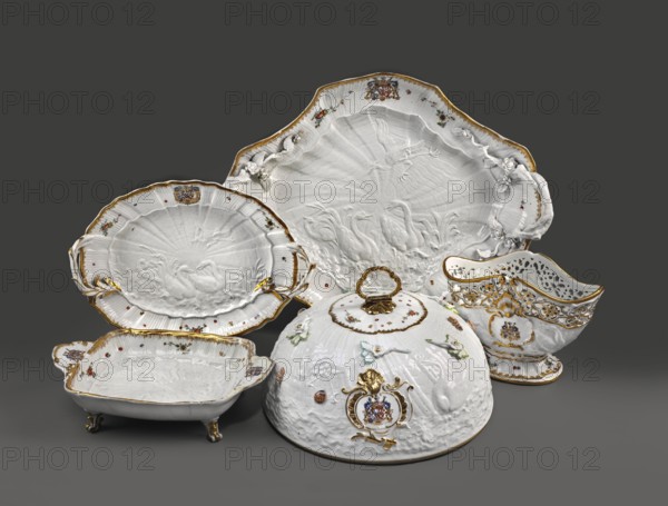 Sweets plate, Kaendler, Johann Joachim 1706-1775 sculptor / author, Koenigliche Porzellain Fabrique, Meissen 1710-1763 manufacturer / author, Eberlein, Johann Friedrich 1695-1749 sculptor / co-creator. Swan Service. Rectangular, on four volute legs, with shell handles on the shorter sides, with the Bruehl marital coat of arms. At the bottom there is a relief with swans, a crane and a heron; the inner sides have a shell relief, Indian flowers and gilding at the edge. 1740. Saxony; Meissen. Vessel. Height 8.5 cm; width 31.0 cm; depth 20.4 cm., A group of ornate white ceramic serving pieces displayed against a dark gray background, including a large scalloped platter, a shallow oval bowl, a footed rectangular dish, a dome-shaped covered dish with a loop handle, and a pierced sauceboat with a pedestal base; the ceramics feature raised molded decoration with motifs of birds and foliage, applied floral details, and gilded trim along rims and handles, with small painted floral accents in colo...