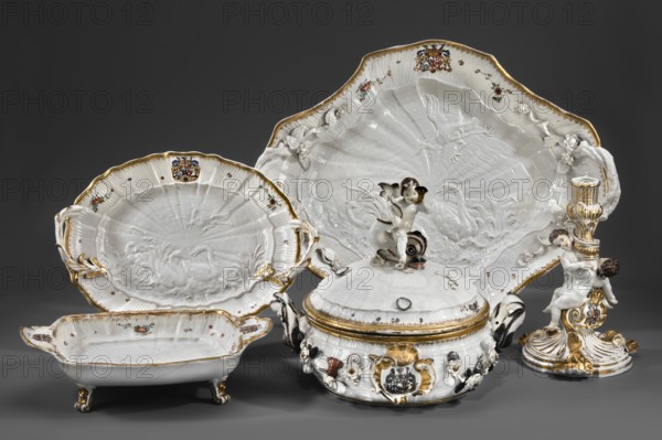 Sweets plate by Johann Joachim Kaendler 1706–1775, modeled/manufactured by Königliche Porzellan-Fabrik Meissen Meissen, Saxony, co-creator Johann Friedrich Eberlein 1695–1749. Swan Service. Rectangular vessel on four volute legs, with shell handles on the shorter sides and the Brühl marital coat of arms. At the bottom a relief with swans, a crane and a heron; inner sides with shell relief, Indian flowers and gilded rim. Porcelain, 1740. Height 8.5 cm; width 31.0 cm; depth 20.4 cm., A group of ornate ceramic serving pieces arranged against a dark gray background including a large scalloped platter, a smaller scalloped dish, a lidded tureen, a rectangular footed bowl, and a decorative candlestick featuring a cherub figure; the ceramics are predominantly white with glossy surfaces and molded relief details, accented with gold trim and highlights, and with small painted floral motifs in colors like red, blue, green, and purple; the tureen has a sculpted finial showing a pair of small figur...