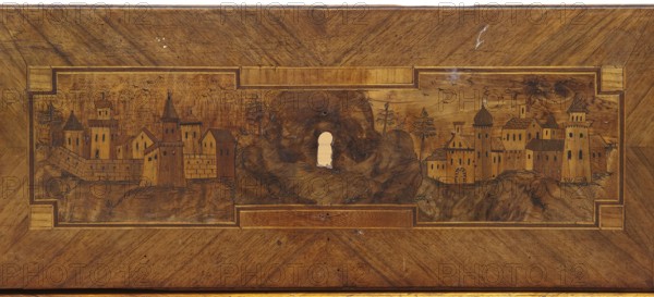 Chest of drawers, provenance revindicated, decorated with roses, classicism style, vedute, geometric ornament, dolphins, eagles, rosettes; ca. 1790; Saxony; piece of furniture, storage piece; height 97 cm, width 143 cm, depth 62 cm, A rectangular wooden panel with a central carved and inlaid scene depicting a stylized townscape set among rocky hills, featuring a group of towers, turrets, domed and pitched roofs, and a few tall trees; the scene is framed by a recessed rectangular border with corner blocks. The wood shows varied brown tones from light golden brown to deeper walnut brown with visible grain patterns and darker burnished lines used to define architectural details and landscape textures; there are a few small scratches and worn spots, and a lighter area where a roughly keyhole-shaped hole appears in the center of the rocky hill.