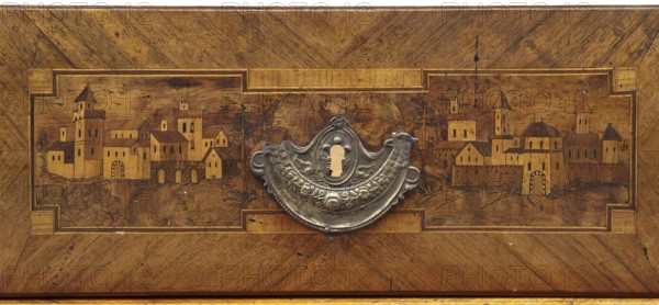 Chest of drawers; creator, revindication provenance; decorated with roses, vedute, dolphins, eagles, rosettes and geometric ornament in a classicism style; ca. 1790; Saxony; piece of furniture, storage piece; height 97 cm, width 143 cm, depth 62 cm, A wooden panel with a rectangular inset showing a stylized town scene carved or inlaid into the wood, featuring a group of buildings with towers, pitched roofs, arches, and small windows; a few trees and undulating ground surround the buildings and a few birds are sketched in the sky. Centered on the panel is an ornate metal escutcheon with a decorative, curved shape and embossed floral and scroll motifs, including a keyhole opening. The wood displays warm brown and golden tones with darker brown grain patterns and some small dark spots and blemishes, and the metal escutcheon appears silver-gray with areas of darker patina.