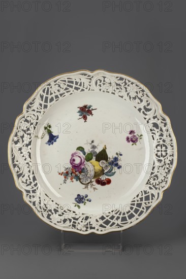 Platter with openwork, Königliche Porzellain-Fabrique, Meissen 1710–1763, ca. 1760. Round platter with a collar with a wavy gilded edge, cut into openwork with a motif of shell volutes and wavy flower branches, divided by two fan-shaped lattices with a flower or palmette motif; relief in the Bruehlsche Allerlei type. In the mirror, a colorful composition with a cut lemon, fig, cherry, currant and drop-shaped flowers, as well as projections of flower branches and a gooseberry branch. Porcelain, Meissen, Saxony. Vessel, height 6.7 cm, diameter 39.6 cm., A round white porcelain plate with a pierced ornate rim featuring floral and lattice cutwork and a thin gold edge, decorated in the center with a painted still life of fruit and flowers including purple, yellow, red, blue, green, and pink tones, with a few additional small floral sprays around the inner rim; the plate sits on a clear stand against a dark gray background.