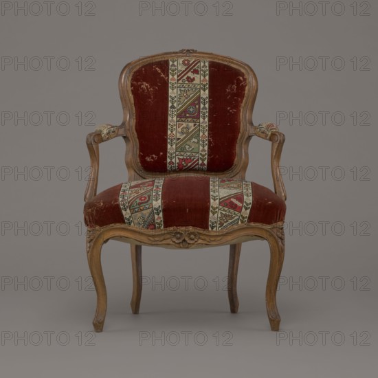 Armchair, rococo, flowers, geometric ornament, ca. 1760, France, piece of furniture seating piece, height 85.5 cm, width 62 cm, depth 60 cm., A vintage wooden armchair with curved armrests and cabriole legs upholstered in deep red velvet with patterned woven fabric panels; the panels feature geometric motifs in beige, green, red, blue, and black, running vertically on the backrest and seat and wrapping over the arm pads; the wood frame is a warm brown with carved floral details on the apron and crest; the velvet shows areas of wear and slight discoloration; the background is a smooth neutral gray.