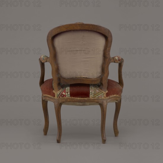 Armchair, rococo style, flowers and geometric ornament, ca. 1760, France; seating piece furniture, height 85.5 cm, width 62 cm, depth 60 cm., A wooden armchair shown from the back with a curved frame and carved top, a pale beige upholstered back panel, wooden curved arms with small padded rests covered in a patterned fabric, and a seat upholstered in worn red fabric with a decorative multi-colored patterned strip; the wooden legs are cabriole style with a natural brown finish, and the background and floor are uniform gray.