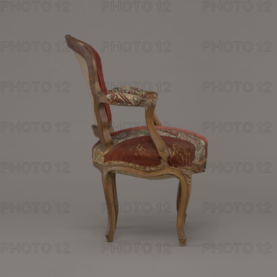 Armchair, rococo, flowers, geometric ornament, ca. 1760, France, piece of furniture seating piece, height 85.5 cm, width 62 cm, depth 60 cm, A wooden chair with curved legs and armrests upholstered in worn red velvet and patterned fabric in a mix of red, green, beige, and black; the wood shows a light brown finish with visible grain and carved details, and the upholstery has areas of discoloration and wear exposing lighter brown patches; the background is a flat gray.