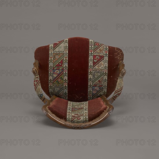 Armchair, rococo, flowers, geometric ornament, ca. 1760, France; piece of furniture seating piece; height 85.5 cm, width 62 cm, depth 60 cm., An upholstered chair viewed from the front showing a curved wooden frame and a fabric back and seat with a striped patterned banding; the visible colors include deep red, beige, green, blue, brown, and hints of yellow and pink, with the wood showing warm brown tones and the background a neutral gray.