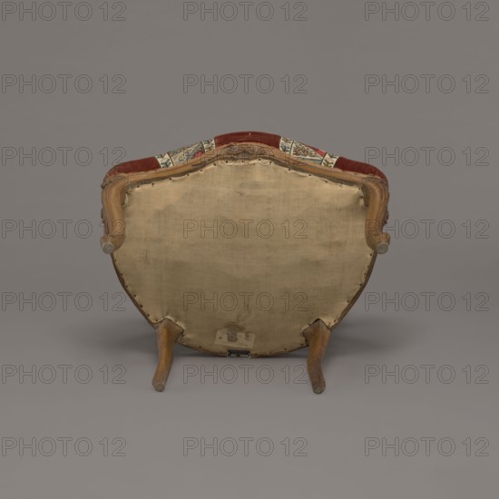 Armchair, rococo, flowers, geometric ornament, ca. 1760, France;, piece of furniture seating piece; height 85.5 cm, width 62 cm, depth 60 cm., An upholstered chair viewed from the back showing a beige fabric panel on the rear, a wooden frame with curved arm supports and legs in a brown wood tone, and a top edge with a strip of red upholstery and patterned fabric in colors including red, green, and cream; the background and floor are shades of gray.