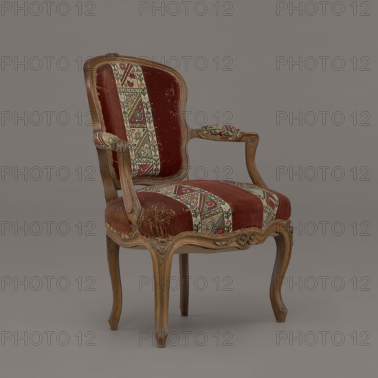 Armchair, rococo, flowers, geometric ornament, ca. 1760, France; piece of furniture seating piece; height 85.5 cm, width 62 cm, depth 60 cm., A wooden armchair with carved curved legs and frame upholstered in a worn red velvet fabric accented by patterned vertical strips in cream, green, red, and black; the armrests have small padded sections covered in the same patterned fabric, and the chair shows scuffs and worn areas on the seat and back; the wood has a light brown finish and the background is a smooth gray.