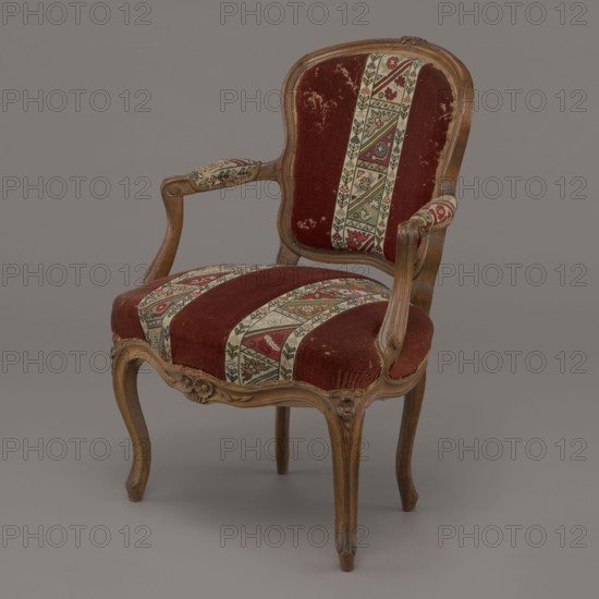 Armchair, rococo, flowers, geometric ornament, ca. 1760, France; piece of furniture seating piece; height 85.5 cm, width 62 cm, depth 60 cm., An upholstered wooden armchair with curved carved legs and a carved crest on the back, showing a worn deep red velvet fabric on the seat and back with patterned vertical bands of beige, green, red, and black tapestry; the wooden frame is a medium brown with visible grain and carved floral details, and the armrests have padded ends covered in the same patterned fabric; the background is a smooth neutral gray.