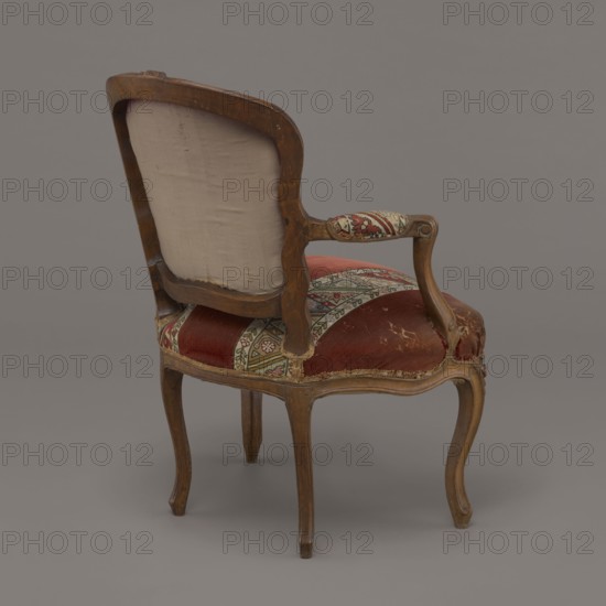 Armchair, rococo style, flowers and geometric ornament, ca. 1760, France; seating piece piece of furniture; height 85.5 cm, width 62 cm, depth 60 cm., An upholstered wooden armchair shown from a rear three-quarter view with a curved back and carved frame, featuring a faded beige fabric on the back panel, patterned upholstery with red and cream fabric on the seat and arm pads, and visible wooden tones of medium brown on the frame and legs against a gray background.