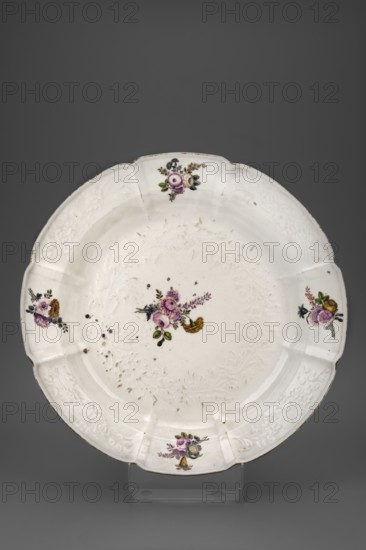 Platter with a Gotzkowski relief, Koenigliche Porzellain Fabrique, Meissen 1710–1763, Gotzkowski relief; Round platter with an octagonal collar with a relief of a flower branch in four fields alternating with plain fields painted with a colorful bouquet of German flowers. In the mirror, within a relief wreath tied with a ribbon, a bouquet with a raspberry rose and a yellow carnation . On the flats of the mirror and collar—small flowers and leaves. Gilded edge heavily worn. c. 1745. Saxony; Meissen. Porcelain vessel; height 7 cm, diameter 39.8 cm., A white ceramic plate with a scalloped rim sits against a gray background, decorated with small floral motifs in purple, pink, yellow, green, and dark blue placed near the center and around the rim; the rim also shows embossed floral patterns in the white ceramic, areas of wear and small dark spots, and a thin band of darker discoloration along parts of the edge; the plate rests on a translucent stand that is visible at the bottom.