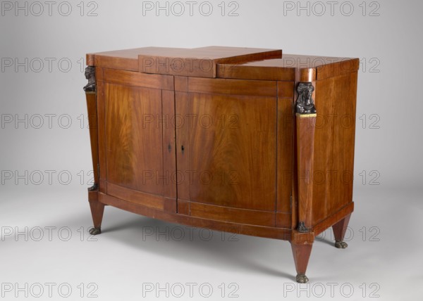 A one-piece cabinet with a slightly rounded front closed by double doors, supported by four low, angular, tapering legs terminating in feet shaped like animal paws. Front corners are chamfered, with herm pilasters. The top has a wide frame, raised in the center and projecting. Entire piece veneered and polished in mahogany. Door leaves have a framed construction with single panels and cubes in the corners; the back is framed with two panels. Female busts, herm feet and leg slopes cast in cast iron. Brass hinges. Empire style, 1815–1825, Warsaw, furniture storage unit. Height 83 cm, width 113 cm, depth 56.3 cm., A wooden cabinet with a warm brown finish stands on tapered legs with dark metallic paw feet, featuring two front doors with rounded panels and a stepped top surface; decorative dark metallic sculpted figures are mounted at the upper front corners above vertical tapered pilasters, and the wood grain displays swirling patterns and subtle variations in brown tones against a neutra...