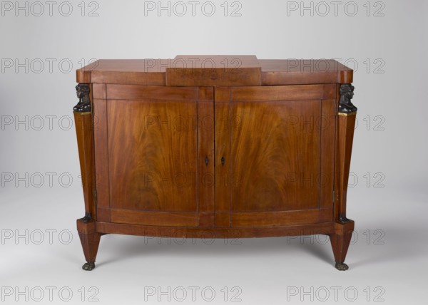 Cabinet, Empire style, mahogany veneered and polished, 1815–1825, Warsaw, one-piece cabinet with slightly rounded front closed by double doors, supported by four low angular tapering legs terminating in animal-paw feet. Front corners are chamfered with herm pilasters; top has a wide frame raised in the center and projecting. Door leaves have framed construction with single panels and corner cubes; back is framed with two panels. Female busts, herm feet and leg slopes cast in cast iron; brass hinges. Function: furniture, storage unit. Dimensions: height 83 cm, width 113 cm, depth 56.3 cm., A wooden cabinet with a warm brown finish and visible wood grain stands against a plain light gray background, featuring a curved front with a pair of central doors and a rectangular top with a stepped center section; each front corner shows a vertical tapered support topped by a small dark sculpted bust and accented with a narrow band of metallic color, and the cabinet rests on tapered legs ending in...