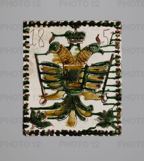 Tile, tiles, eagles, stoves, plant decorations, Hutsulshchyna, Decoration with a double-headed eagle motif, 1851, Hutsulshchyna, tile, height 20 cm, width 23 cm, depth 6 cm, A rectangular ceramic tile with a cream background framed by a border of alternating dark green and brown dots shows a stylized double-headed bird motif in green and mustard yellow with brown outlines; the bird has two curved heads with beaks, textured scale-like markings on the necks, a central body with a basket-like pattern, layered wings with green and yellow feathers, and a fan-shaped tail with green and yellow elements; each wing is flanked by a long brown sword-like object held by a yellow limb; there are decorative green floral shapes near the lower corners and horizontal green bands behind the central figure.
