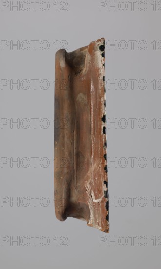 Tile, ceramic tile with eagles and plant decorations, Hutsulshchyna, Decoration with a double-headed eagle motif, 1851; tile, height 20 cm, width 23 cm, depth 6 cm, The image shows a vertically oriented fragment of a terracotta object suspended against a plain light gray background; the fragment is mostly reddish-brown with areas of darker brown and patches of pale beige or off-white residue, and there are several small irregular black spots along one narrow edge; the surface appears weathered with rough texture, smudges, and some gloss in places, and the shape includes a concave channel and a rounded protrusion at one end.