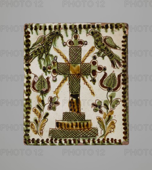Tile, ceramic tiles with plant decorations and Maltese crosses, depiction of two birds, Hutsul region, 19th century, tile, height 20 cm, width 23 cm, depth 6 cm, possibly used for stoves; decoration with a cross motif., A square ceramic tile with a cream background framed by a decorative border in brown and green features a central ornate cross rendered with crosshatched and solid segments in brown and green atop a stepped base; flanking the cross are stylized floral motifs with tulip-like flowers and leafy stems in green and brown, and a pair of birds perched near the top corners with green and brown plumage and dark outlined details; small ornamental dots and curving lines in brown and green accent the design, and touches of yellowish-brown appear in shaded areas and highlights.