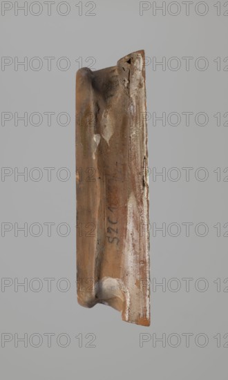 Tile; maker, ceramic tiles; depiction of two birds; plant decorations; Maltese crosses and other crosses; decoration with a cross motif; Hutsul region; 19th century; tile; height 20 cm, width 23 cm, depth 6 cm; used for stoves., A single elongated object with an irregular, worn surface suspended against a plain gray background, showing a dominant brown color with areas of lighter tan and off-white patches, along with small traces of darker brown and faint blackish marks; the object has curved and straight edges, a notch at one end, textured rough spots, and a few smudged lighter streaks and spots.