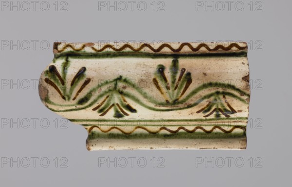 Cornice tile fragments 4, tile cullet; painted with a wavy twirl pattern; 19th century; Hutsul region, Poland; plant decorations; used in stoves., A ceramic fragment with a curved profile showing a cream background with visible crazing and worn edges, decorated with green and brown painted motifs including wavy lines and stylized fan- or leaf-like shapes, and bands of zigzag patterns near the top and bottom.