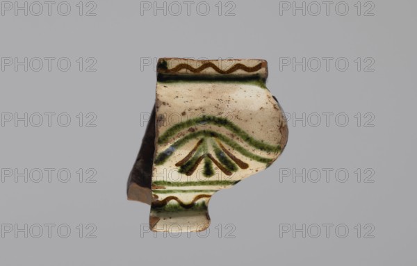 Cornice tile fragments 4. Tile cullet; painted with a wavy twirl pattern; 19th century; Hutsul region, Poland; plant decorations., A fragment of a ceramic object with an off-white glazed surface decorated with painted wavy and arched motifs in green and brown, showing a curved profile with a broken edge and some areas of exposed brown clay, small dark speckles on the glaze, and a neutral gray background.