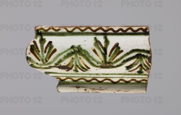 Cornice tile fragments 4, tile cullet; painted with a wavy twirl pattern; 19th century; Hutsul region, Poland; plant decorations; made for stoves., A fragment of a decorative ceramic tile with a white background showing a painted pattern in green and brown; the fragment has a curved molded edge and a scalloped top border in brown, with a repeating wavy brown line and green wash beneath it, and a central leafy or fan-like motif rendered with green brushstrokes and brown accents spread across the surface, with worn edges and small areas of exposed clay in a tan color, all set against a neutral gray backdrop.
