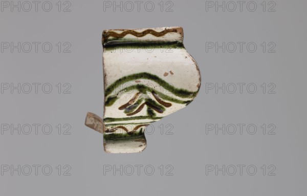 Cornice tile fragments 4, painted with a wavy twirl pattern; tile cullet; stoves; plant decorations; 19th century; Hutsul region, Poland., A fragment of a glazed ceramic object with an off-white base color and visible areas of green and brown glaze, showing curved and wavy painted lines and a small cluster of curved brushstrokes in green and brown, with some exposed unglazed clay at the edges and small chips, photographed against a uniform gray background.